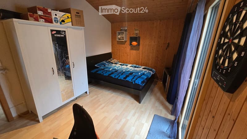 Bedroom with wooden walls and floor, a double bed with blue and black bedding, a large white wardrobe with mirrored doors, a dartboard on the wall, and a small shelf with decorative items.