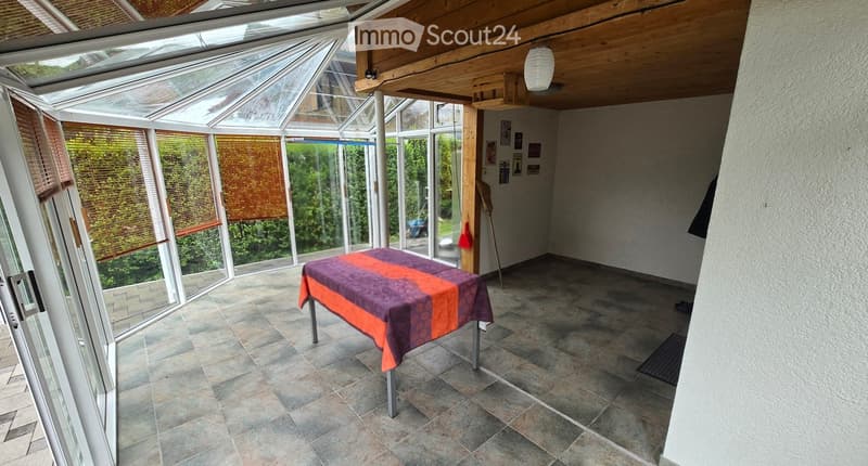 Bright conservatory with tiled floor, large windows, and a single table with a colorful tablecloth.