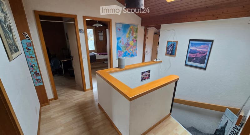 Bright hallway with wooden flooring, white walls, and a wooden railing leading to a bedroom area. Framed pictures and a small shelf with a speaker are visible.