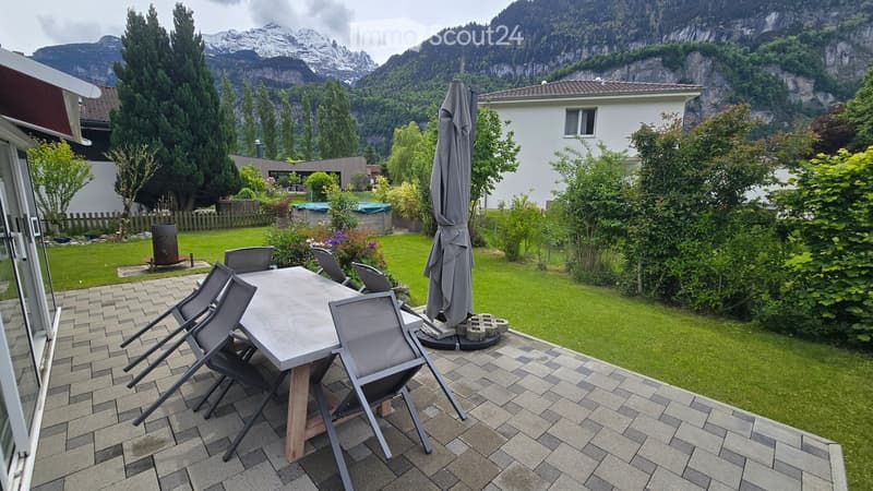 Outdoor patio with a wooden table, four chairs, and an umbrella, surrounded by a well-maintained lawn and trees, with a scenic mountain view in the background.