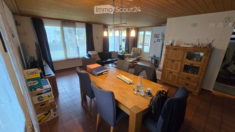 A well-lit living and dining area with a wooden dining table, chairs, a sofa, and a cabinet. The space features large windows with curtains and a hanging light fixture.
