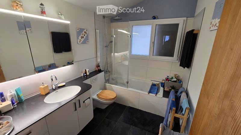 Modern bathroom with a large sink, toilet, bathtub with a glass shower enclosure, and a window. The space includes a towel rack with towels, a stool, and various toiletries on the counter.