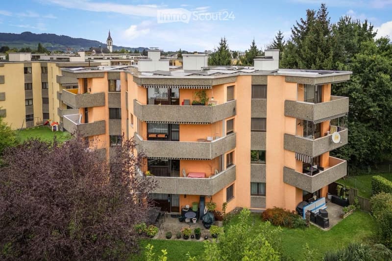 Multi-story residential building with balconies, surrounded by greenery and a view of a town with a church tower in the background.