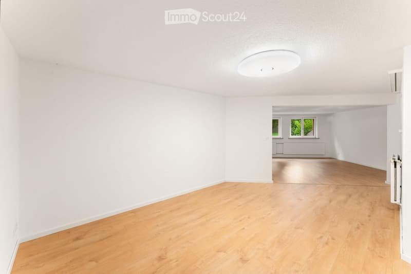 Empty room with light wooden flooring and white walls, featuring a recessed ceiling light and a doorway leading to another room with windows.