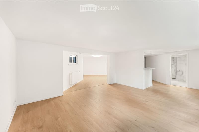 Empty room with light wooden flooring, white walls, and a small window. A radiator is visible on one wall, and a doorway leads to another area.