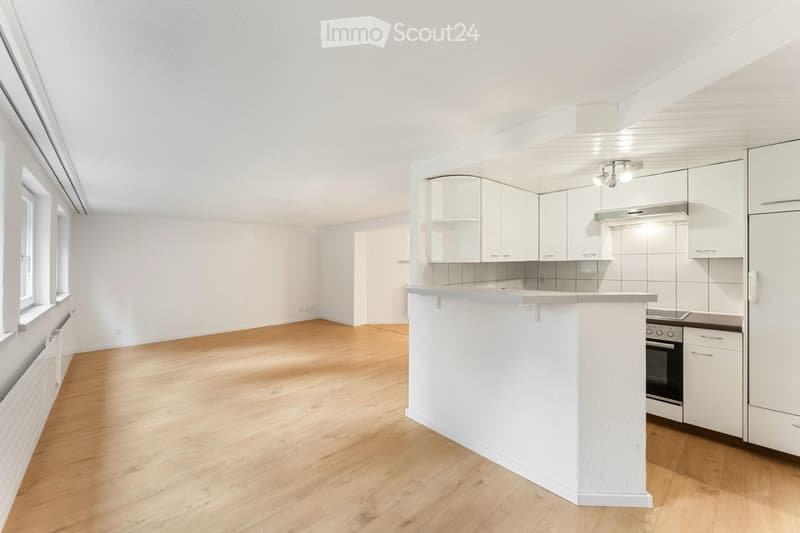 Bright, open-plan room with light wooden flooring, white walls, and large windows. The space includes a kitchen area with white cabinets, a countertop, and built-in appliances, but no furniture or personal items are present.