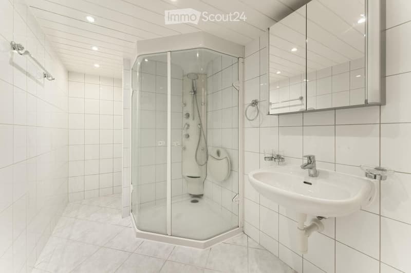 Modern bathroom with white tiled walls and floor, featuring a glass-enclosed shower and a white sink with a mirror above it.