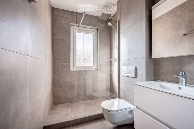 Modern bathroom with grey tiled walls, a white toilet, a sink with a modern faucet, a shower stall with a glass enclosure, and a window for natural light.