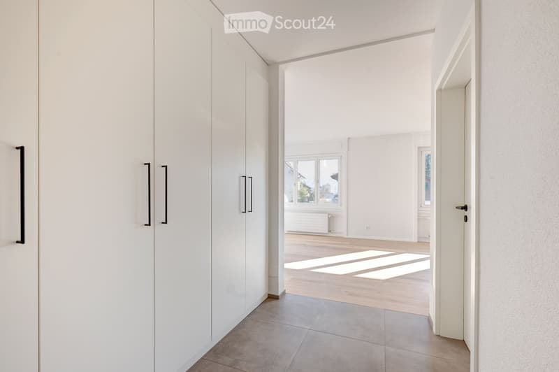 Bright, minimalist hallway with white walls, wooden flooring, and large windows allowing natural light to flood the space. Features modern white cabinetry on one side and a tiled floor in the foreground.