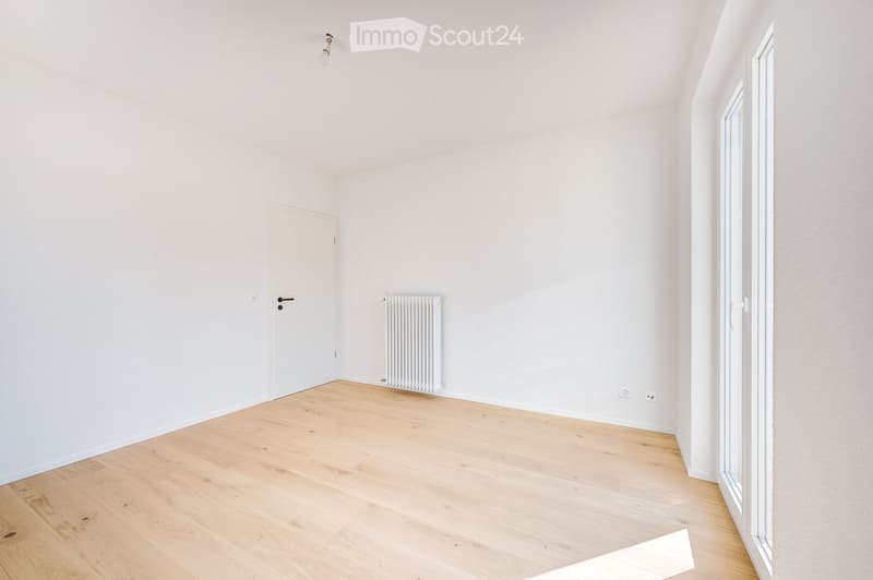 Empty room with light wooden flooring, white walls, a white door, a radiator, and a window with white frame.