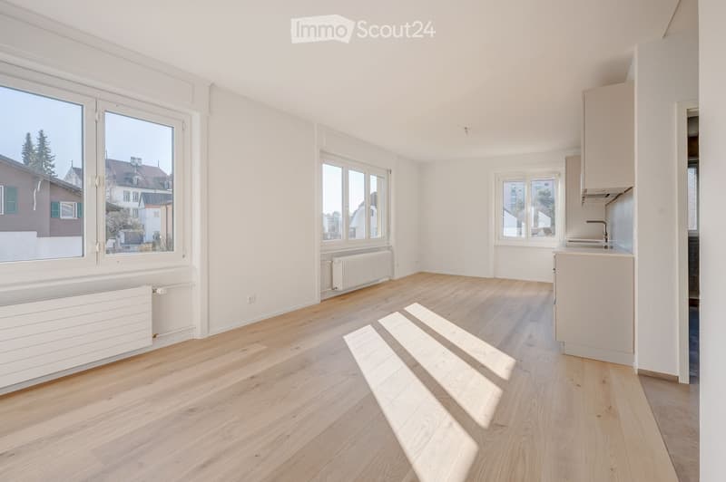 Empty room with light wooden flooring, white walls, large windows, and a small kitchen area with a sink and cabinets.