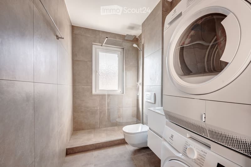 Modern bathroom with a toilet, washing machine, and a glass-enclosed shower area. The space features beige tiles and a large window providing natural light.