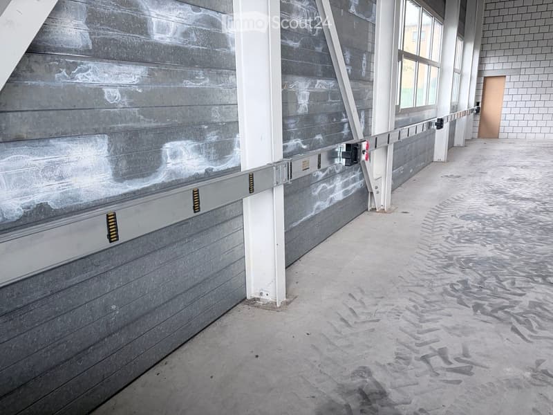 Empty hallway with concrete walls, metal railings, and a tiled floor. The walls have a textured finish and some markings, and there is a door visible at the end of the hallway.