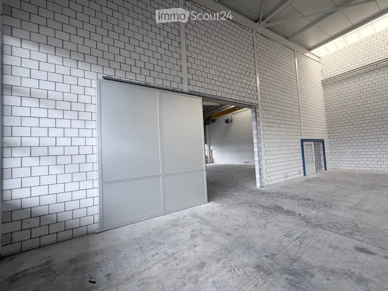 Empty industrial-style room with white brick walls, concrete floor, large garage door, and a small blue-framed doorway leading to another space.