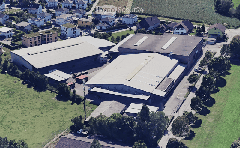 Aerial view of a residential and industrial area with multiple buildings, including houses, warehouses, and a large industrial complex with a prominent roof structure. The area is surrounded by green fields and trees.