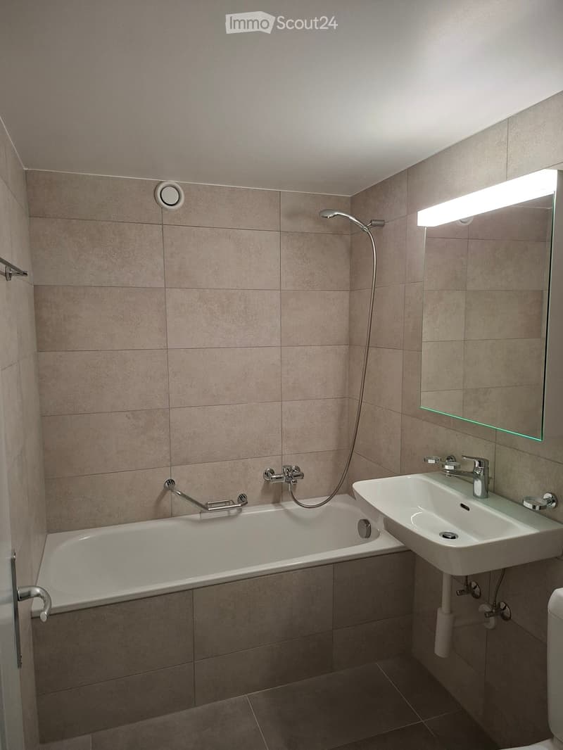 Modern bathroom with beige tiled walls, a bathtub with a handheld shower, a sink with a modern faucet, and a large mirror with built-in lighting.