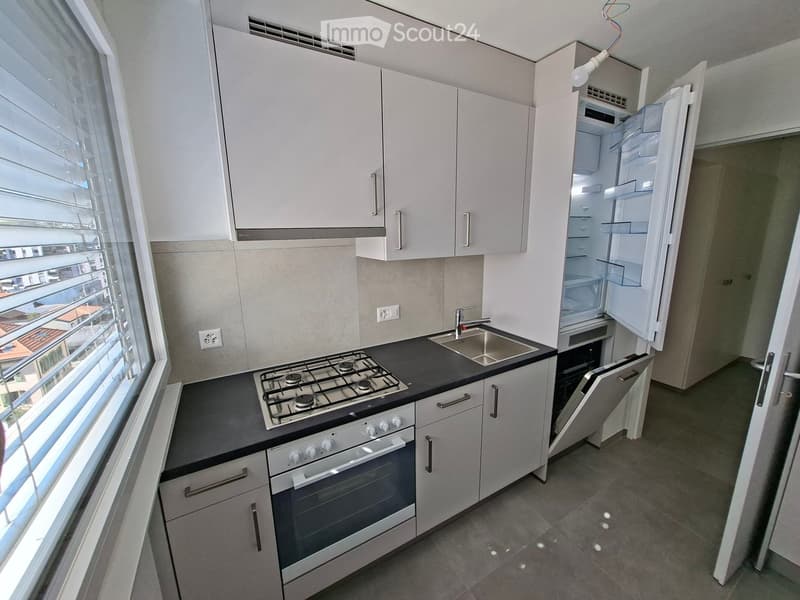 Modern kitchen with white cabinets, gas stove, oven, refrigerator, and sink. Large window provides natural light.