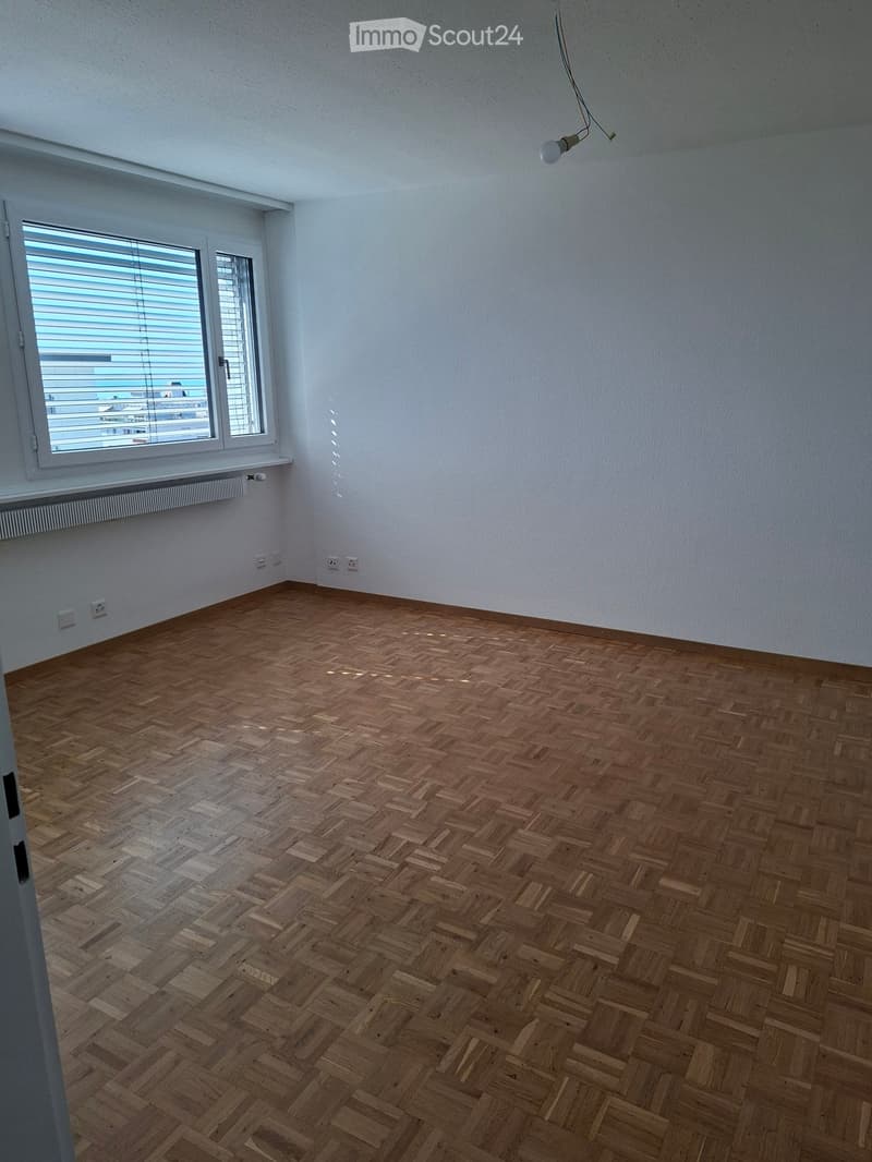 Empty room with wooden herringbone flooring, white walls, and a large window with blinds. No furniture or equipment is present.