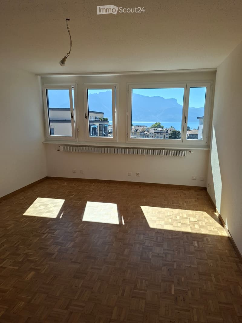 Empty room with wooden herringbone flooring, white walls, large windows offering scenic views, and a single ceiling light fixture.