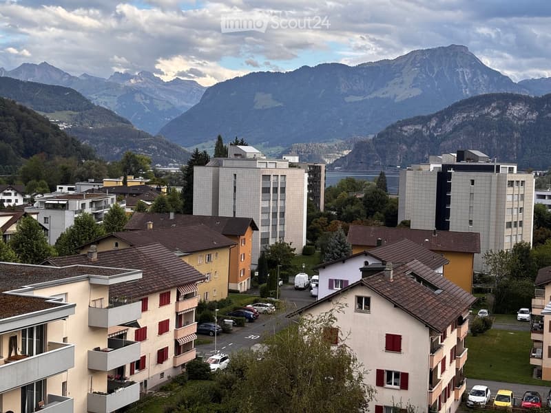 A panoramic view of a town with multi-story residential buildings, surrounded by lush greenery and distant mountain ranges.