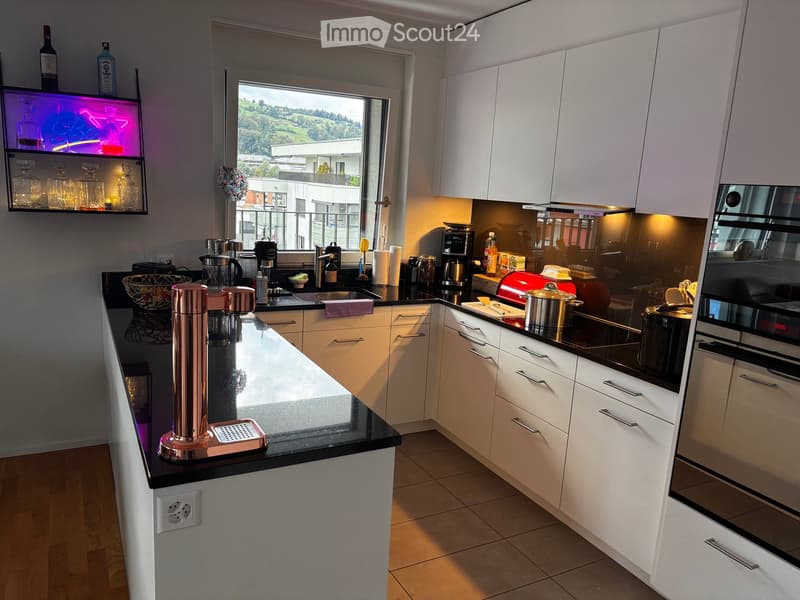 Modern kitchen with white cabinets, black countertops, and various appliances including a coffee machine, oven, and fridge. A large window offers a scenic view of the surroundings.