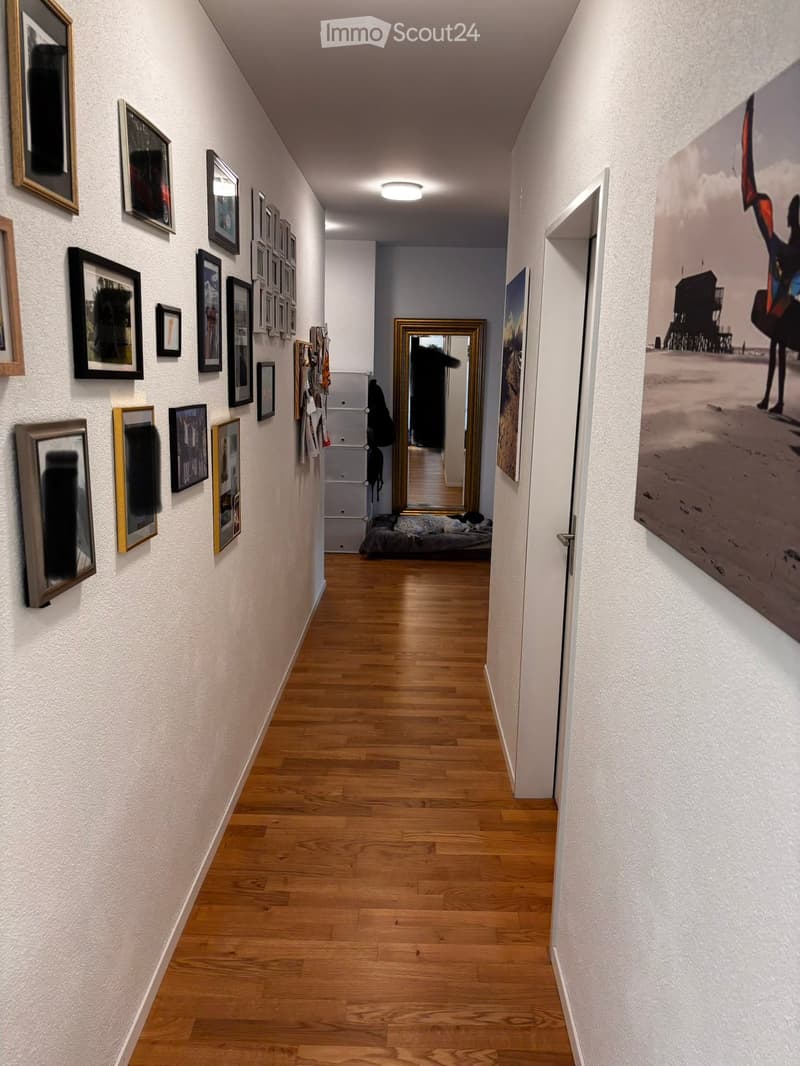 A long, narrow hallway with wooden flooring and white walls, decorated with multiple framed pictures and a large mirror at the end.