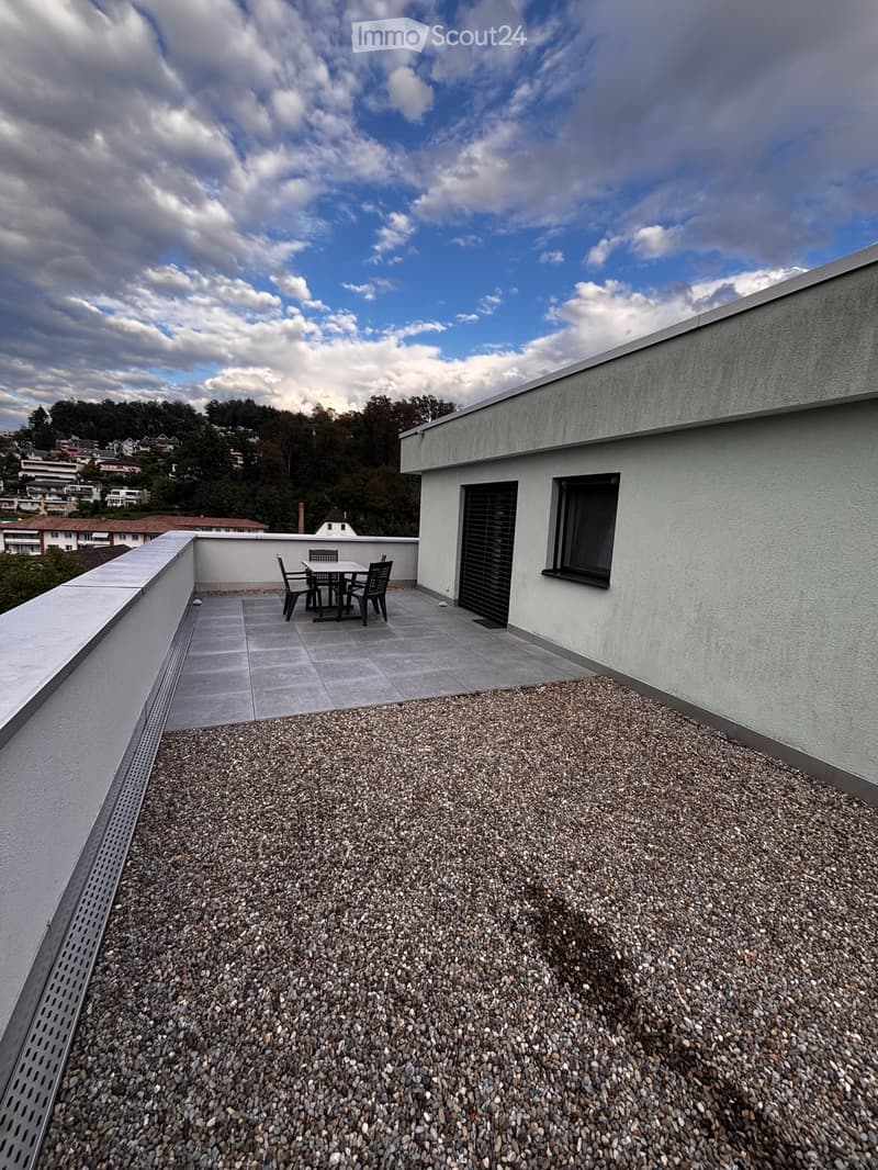 Roof terrace with a small seating area featuring a table and chairs, surrounded by a gravel surface and modern building architecture.