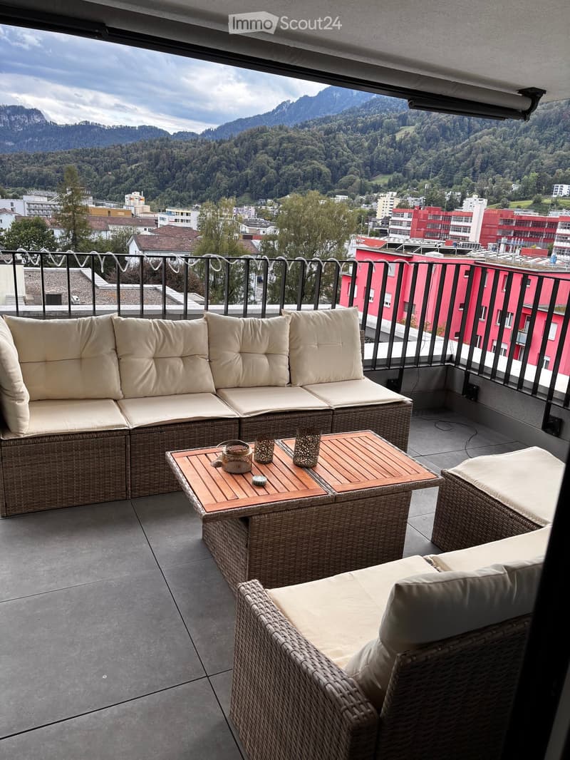 Balcony with wicker furniture, including a sofa, a table, and a chair, overlooking a scenic mountain and city view.