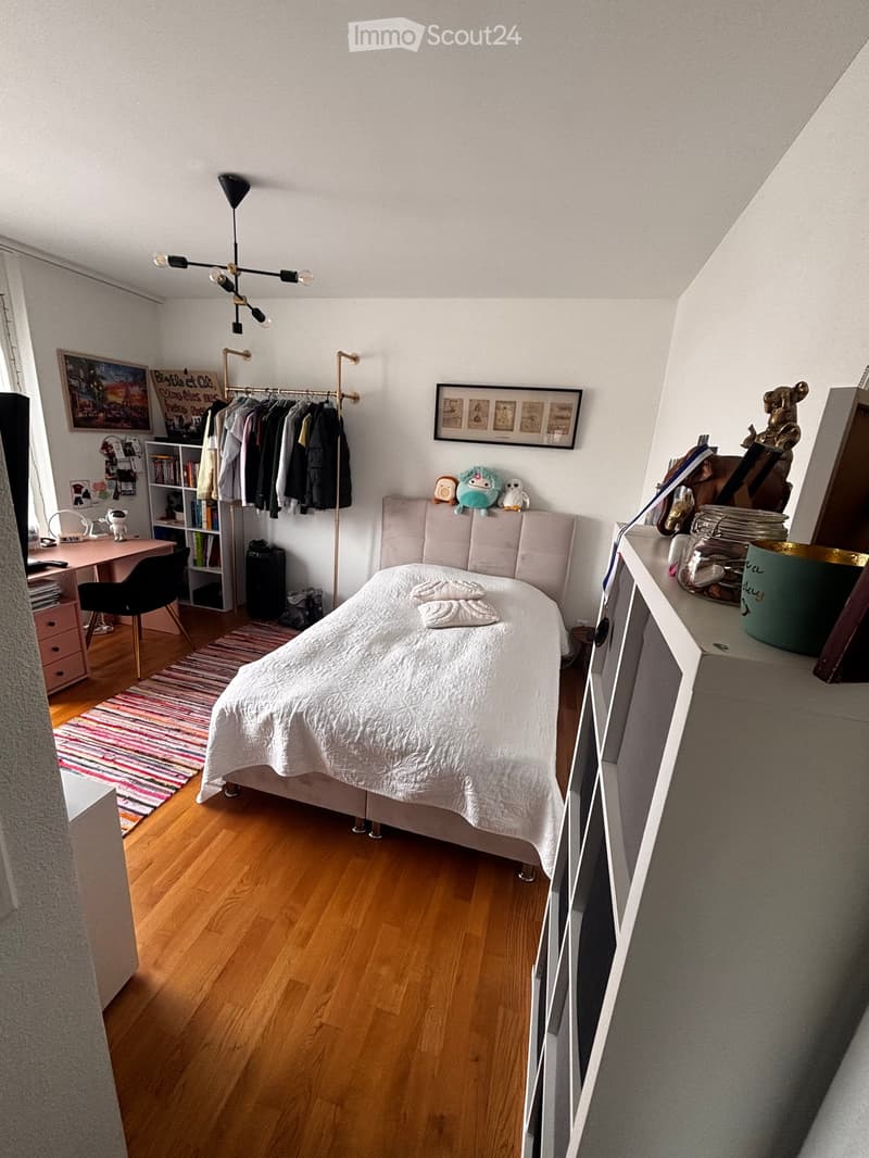 Cozy bedroom with a wooden floor, a neatly made bed, a desk with a chair, a bookshelf, a wardrobe, and decorative items including framed pictures and a rug.