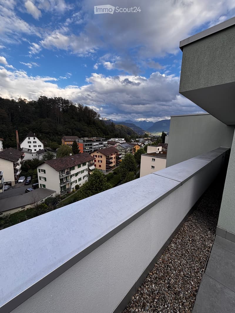 Balcony with a view of a town, surrounded by trees and mountains under a partly cloudy sky.