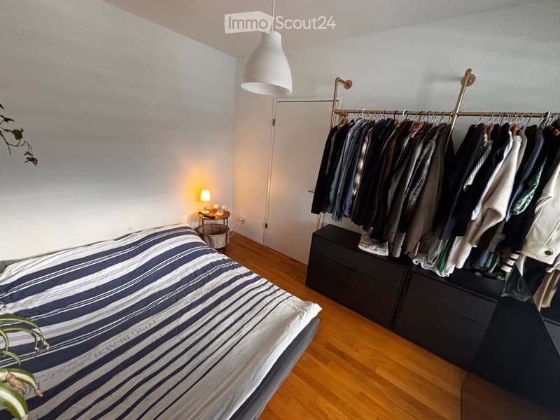 A modern bedroom with a neatly made bed, a wardrobe with hanging clothes, a small table with a lamp, and a hanging plant.