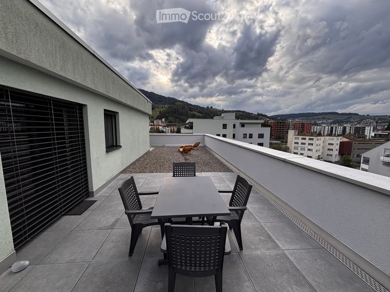 Balcony with a table and four chairs, overlooking a cityscape with buildings and hills under a partly cloudy sky.