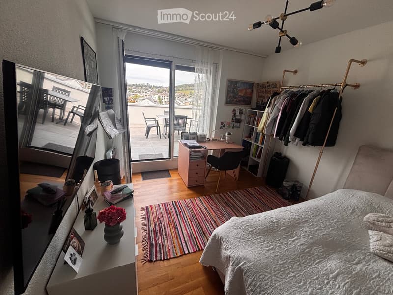 Cozy bedroom with a bed, desk, and large window leading to a balcony. Features a colorful rug, mirror, and decorative items on a side table.
