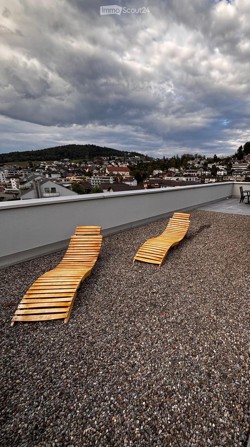 Roof terrace with two wooden lounge chairs and a small table, overlooking a town with houses and hills under a cloudy sky.