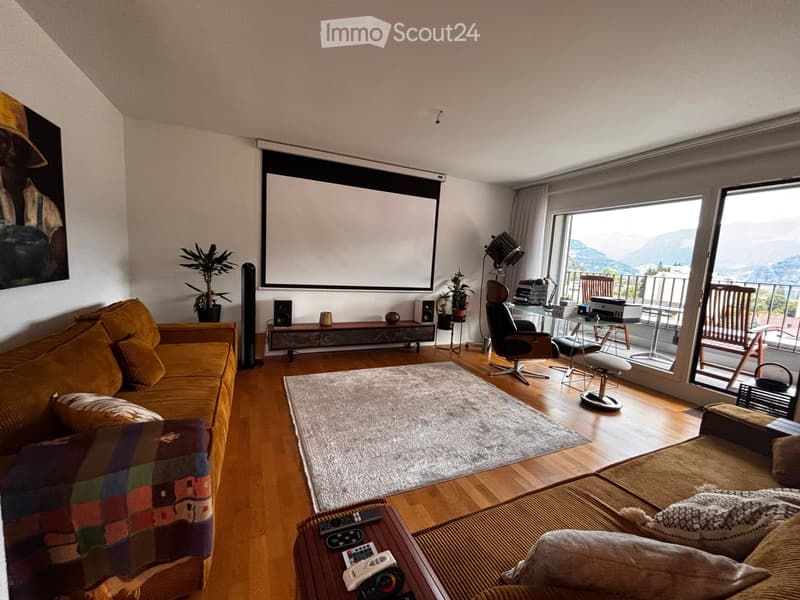 Modern living room with wooden flooring, large windows offering a scenic view, a projector screen, a comfortable sofa, a coffee table, and a small workspace area with a chair and desk.