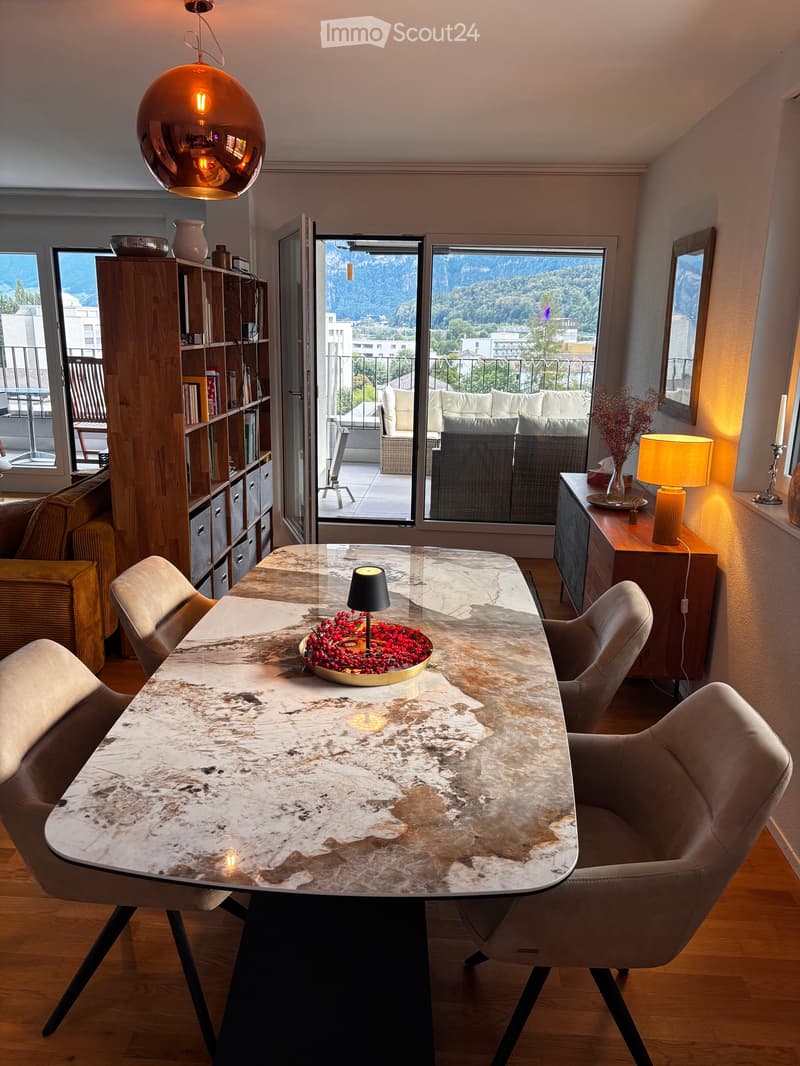 Modern living room with a large marble dining table, chairs, sofa, bookshelf, and a view of the mountains through large windows.