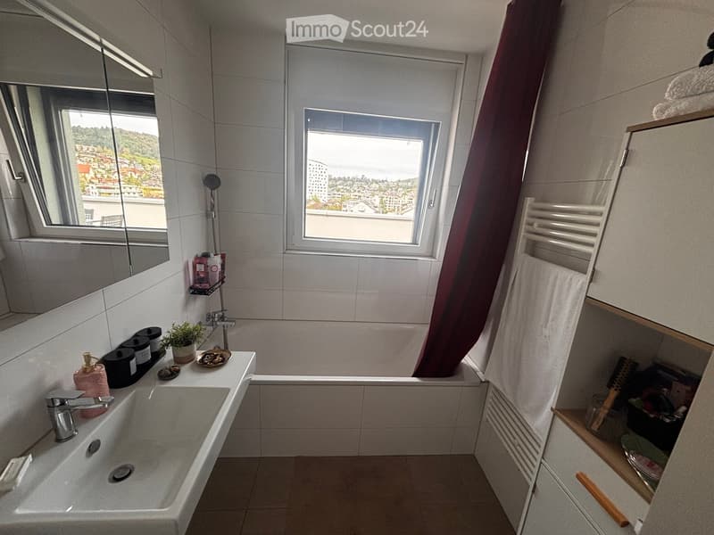 Modern bathroom with a large window offering a city view, featuring a sink, bathtub, mirror, towel, and cabinet.