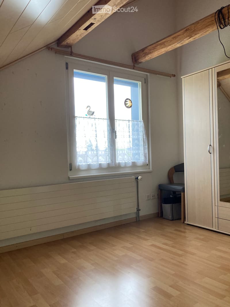 Empty attic room with wooden floor, slanted wooden ceiling beams, large window with white curtains, radiator, and a small cabinet with a mirror.