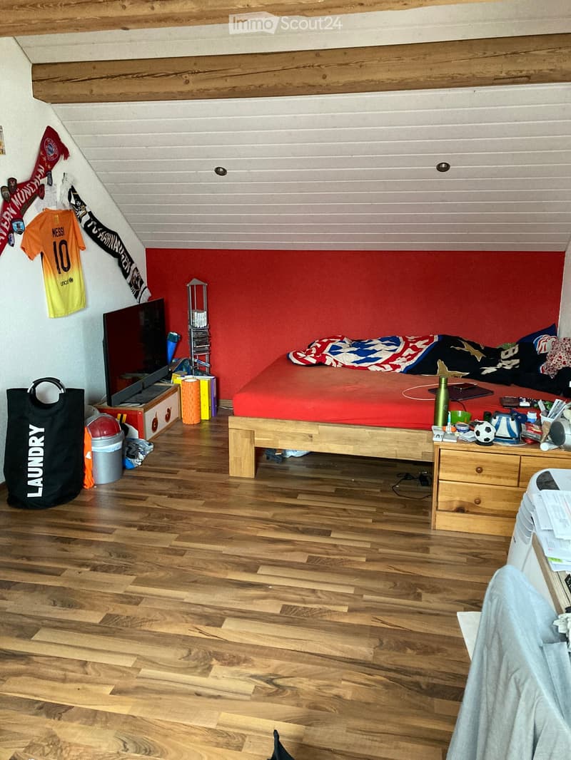 A cozy bedroom with wooden flooring, a bed with red bedding, a laptop on a wooden table, a TV screen, and various personal items including a soccer ball, books, and a trash can.