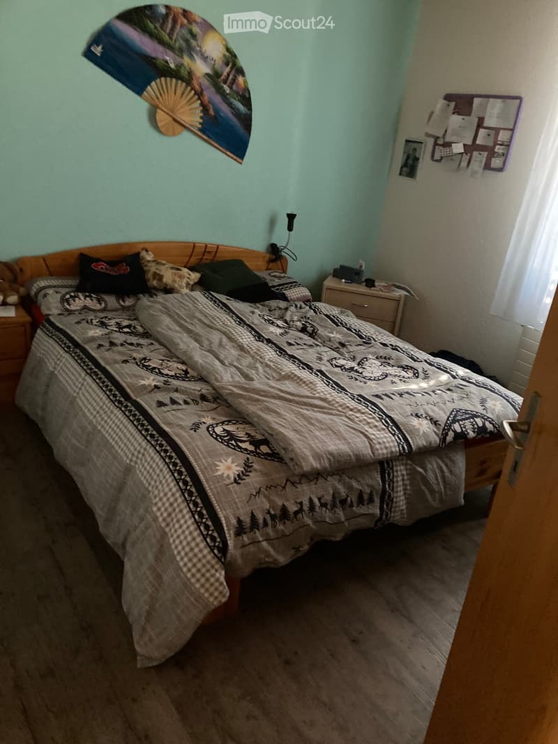 Cozy bedroom with a wooden bed, patterned quilt, nightstand, and wall decorations including a fan and picture frames.
