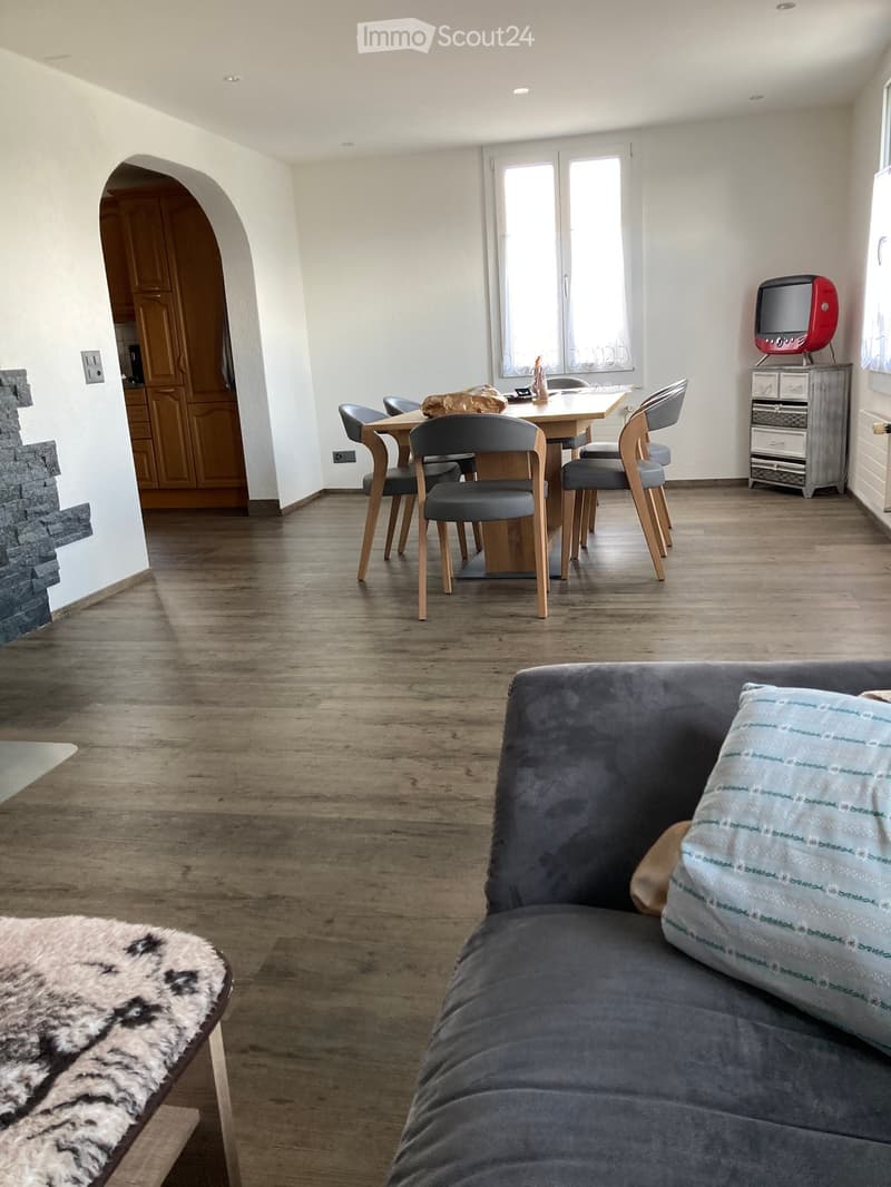 A modern living room with a grey sofa, wooden dining table, six chairs, a small television on a cabinet, and a fur throw on a bench. The room features wooden flooring and white walls.