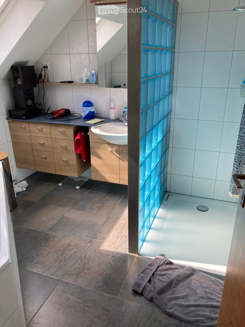Modern bathroom with a sink, mirror, wooden cabinet, and a glass-enclosed shower area. The floor is tiled, and there is a towel on the floor near the shower.