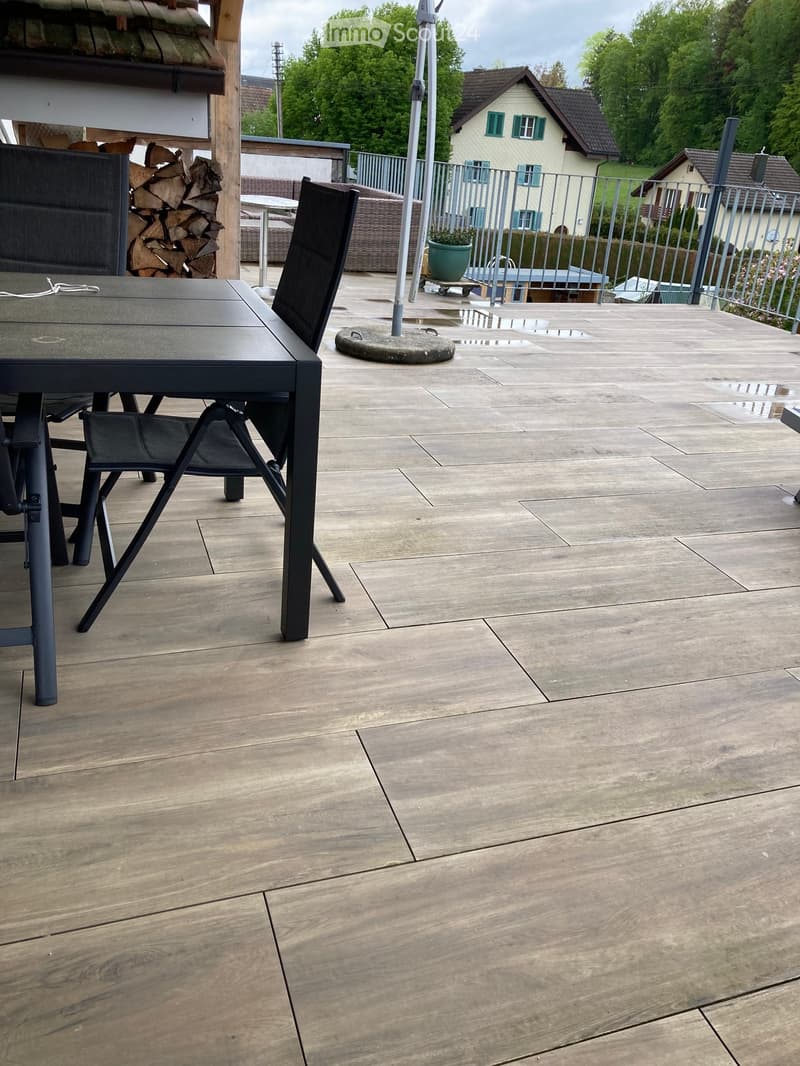 Outdoor patio with a stone floor, a black metal table and chairs, a stack of firewood, and a view of a house and greenery.