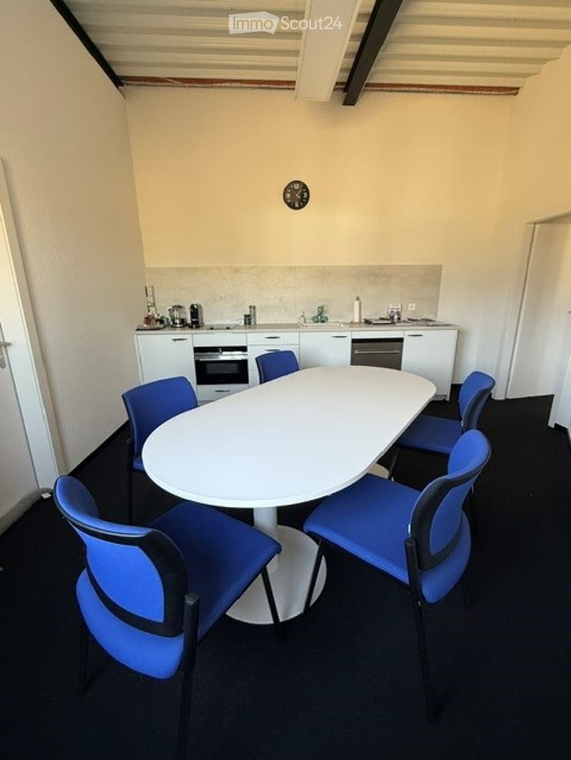 A modern, well-lit room with a white table surrounded by blue chairs, a small kitchen area with white cabinets, an oven, and a dishwasher.