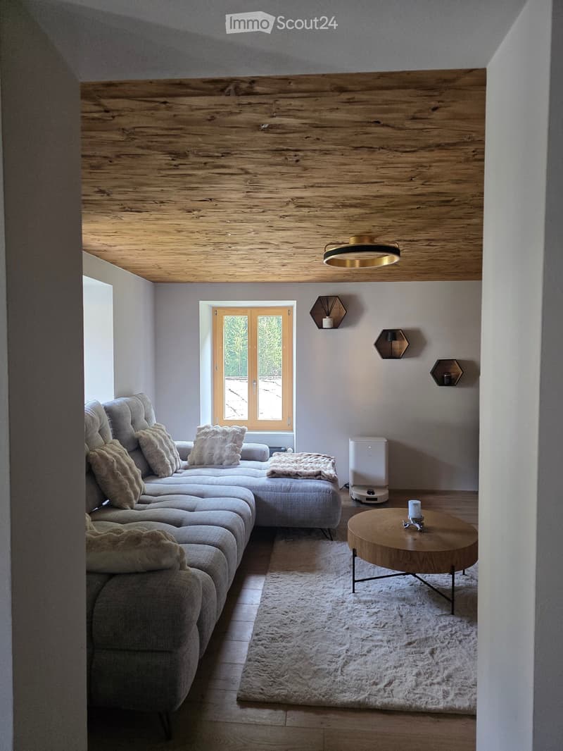 Modern living room with a grey sectional sofa, wooden coffee table, and wall-mounted candle holders. The room features a wooden ceiling and a large window with a view of the outdoors.