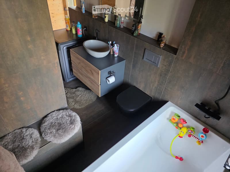Modern bathroom with a dark grey cabinet, white sink, black toilet, and bathtub with toys. Includes a mirror, toiletries, and a furry rug.