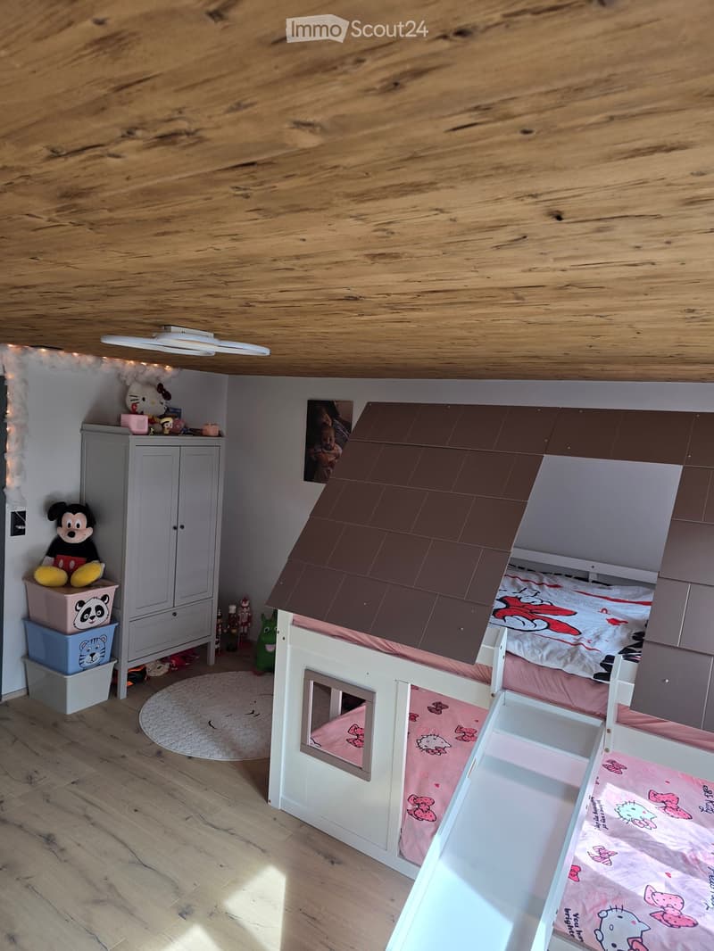 A cozy children's bedroom with a lofted bed designed like a house, featuring a pink Hello Kitty bedding, a wardrobe, and various toys including a Mickey Mouse plush and a panda toy.