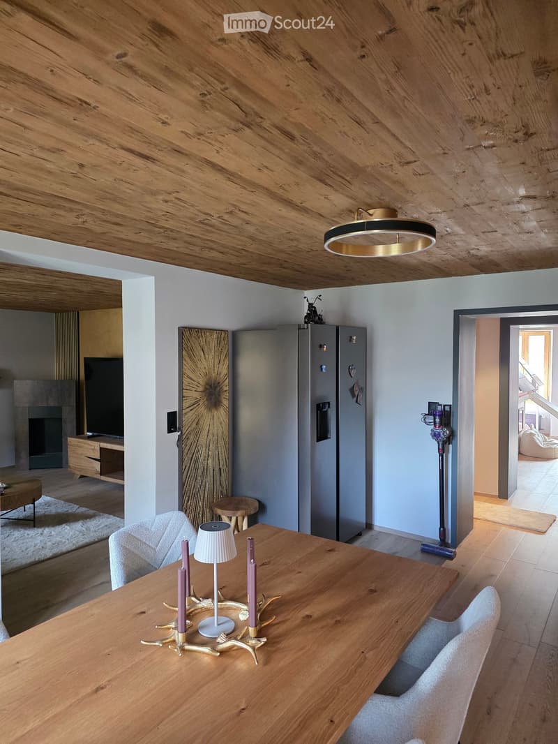 Modern living and dining area with wooden ceiling, white walls, and light wood flooring. Features a dining table with chairs, a lamp, a refrigerator, a television, and a fireplace.