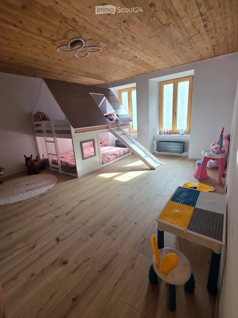 Bright, wooden-floored bedroom with a bunk bed featuring a slide, a small table with toys, and a radiator under a large window.