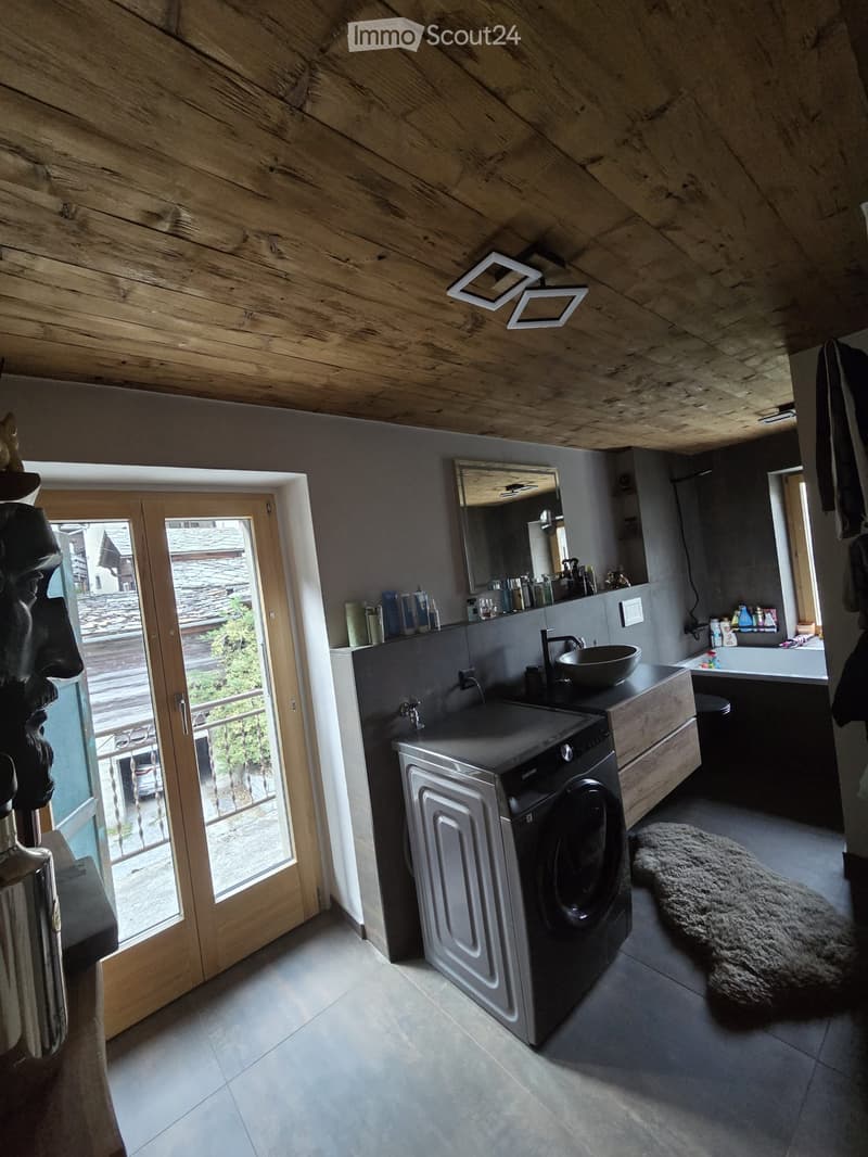 Modern bathroom with wooden slanted ceiling, grey tiles, sink, mirror, washing machine, bathtub, and a view through a large window.